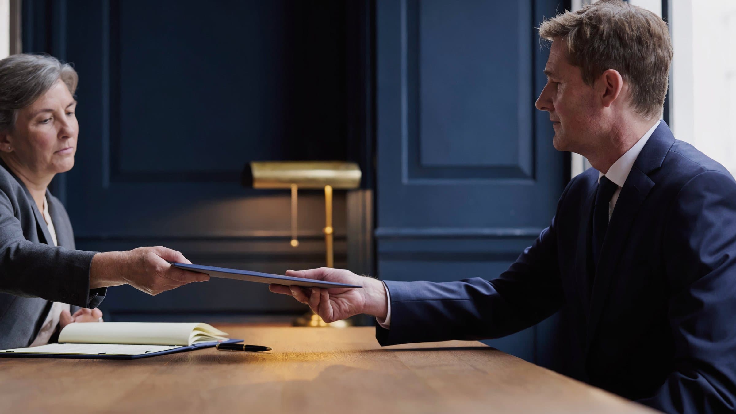 A trust officer handing a document folder across a table to an auditor.