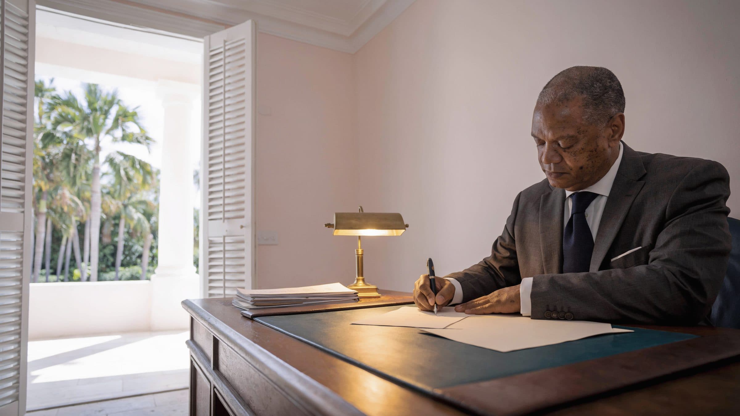 A trust officer signing a page at a desk in a pale pink room, tall shutters open to a courtyard of palms.