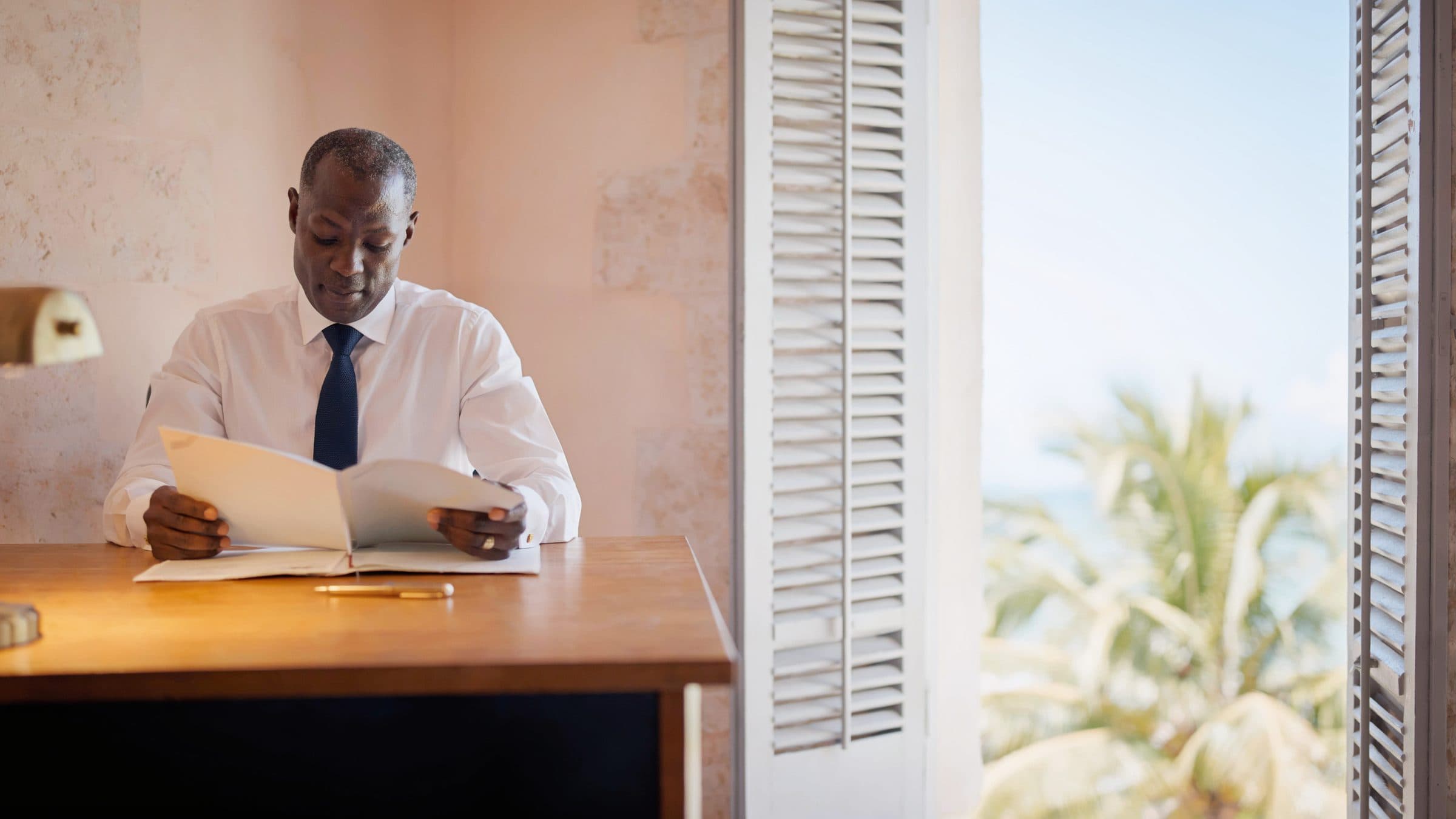 A compliance officer reading a client file at a desk in a room of pale coral stone, shutters open to palms.