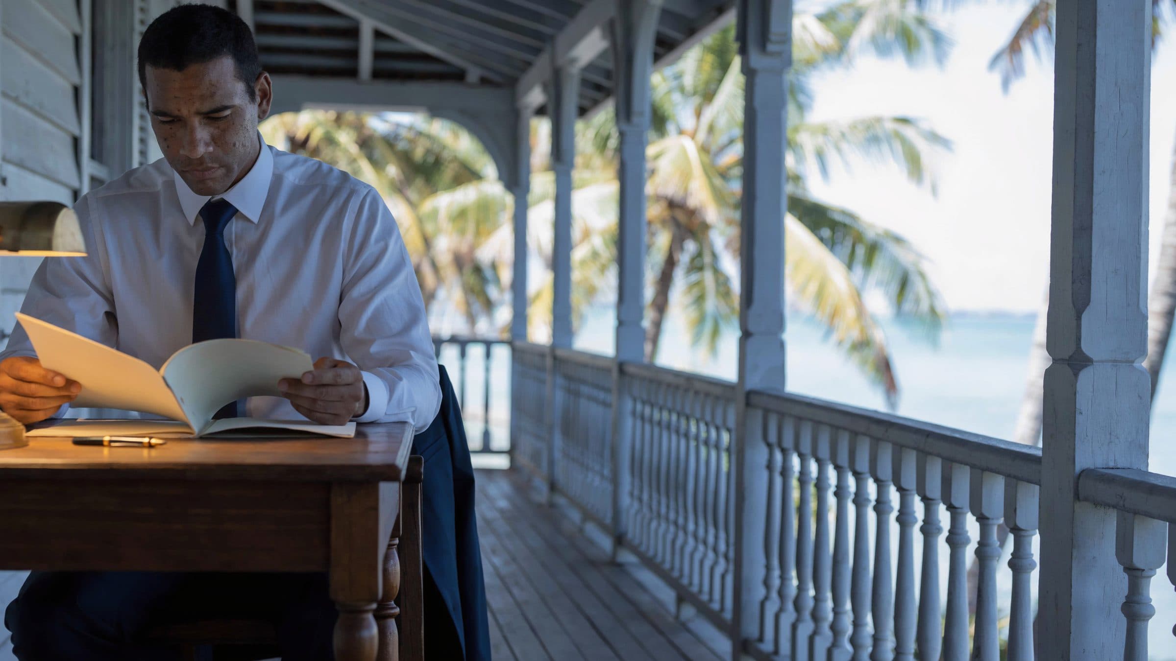 A compliance officer reading a client file at a table on the verandah of a white timber building, palms and the sea beyond.