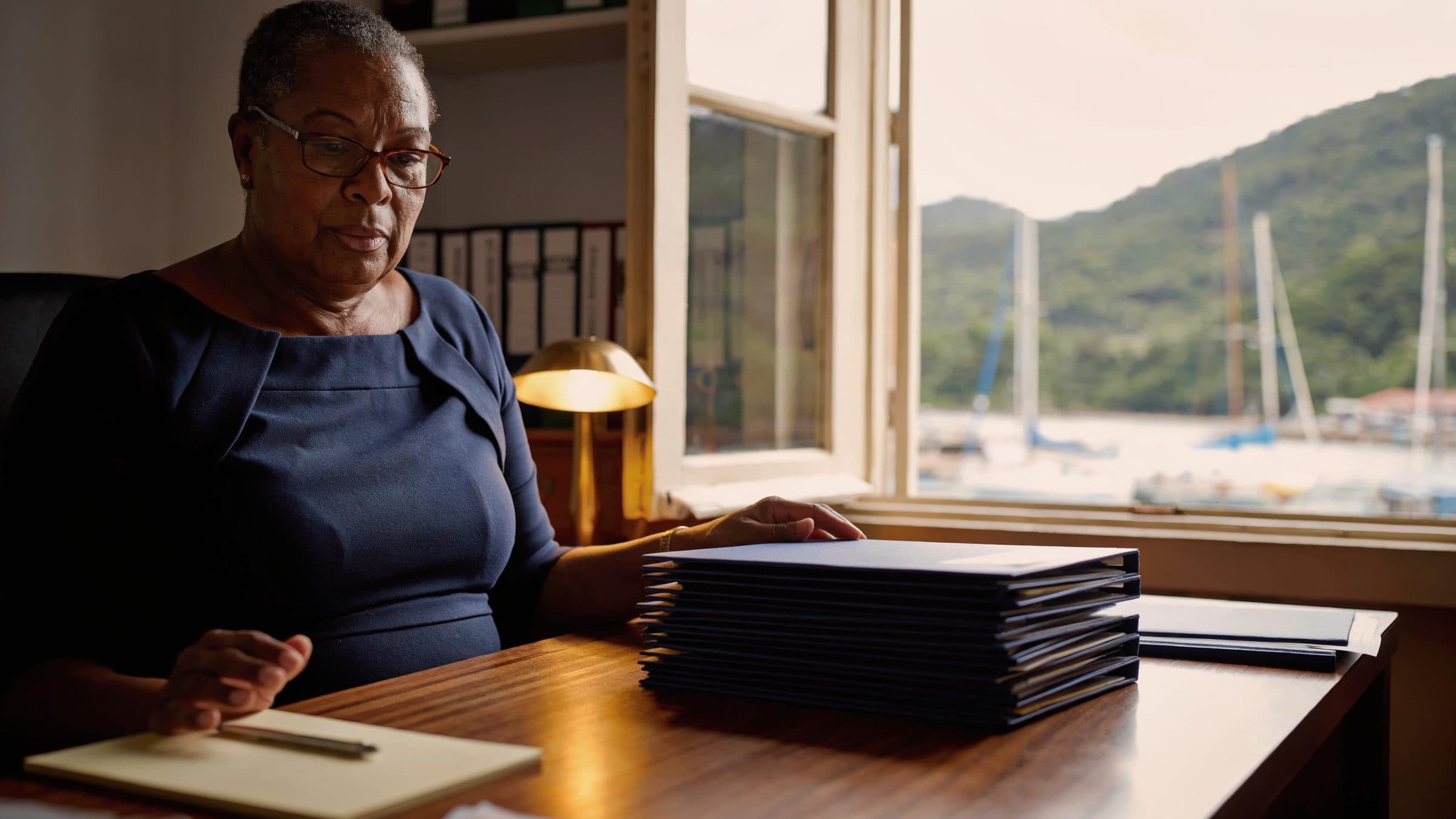A registered agent at her desk with a stack of client files, a harbour of moored sailing boats through the window.