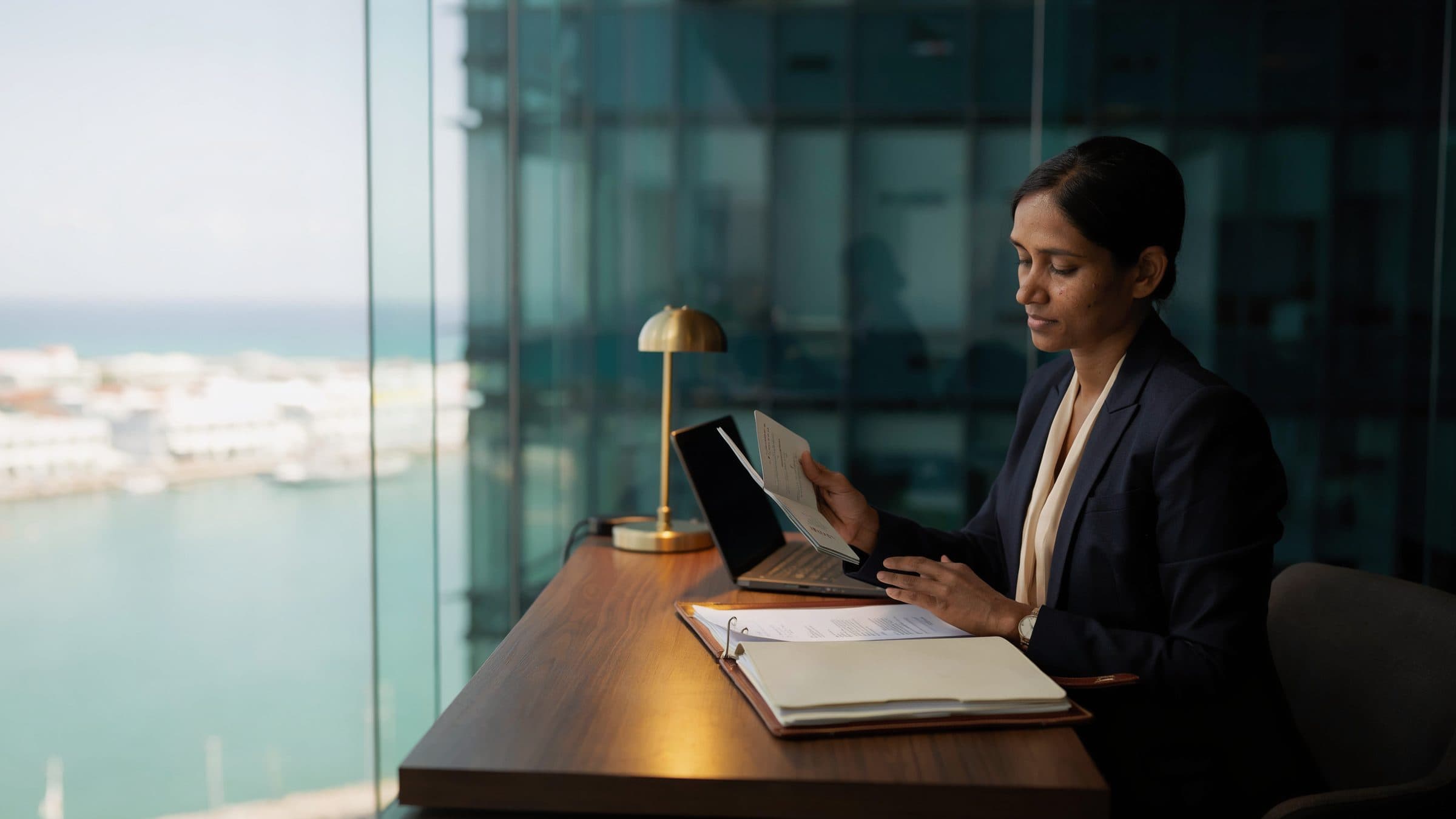 A compliance officer at a desk in a glass-walled office above a harbour, holding a passport beside an open client file.