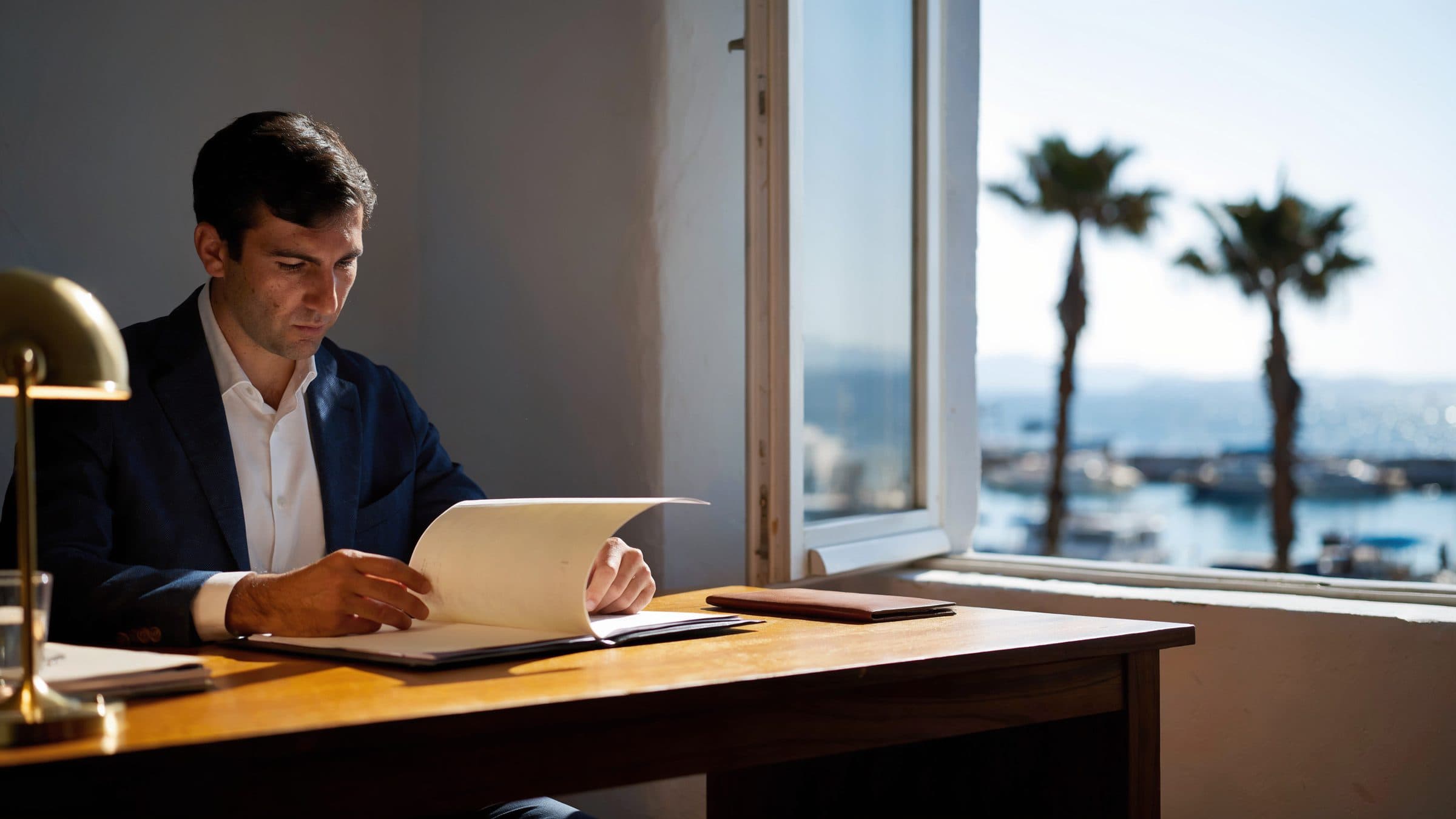A compliance officer reading a client file at a desk in a white-walled office, a harbour and palms through the window.