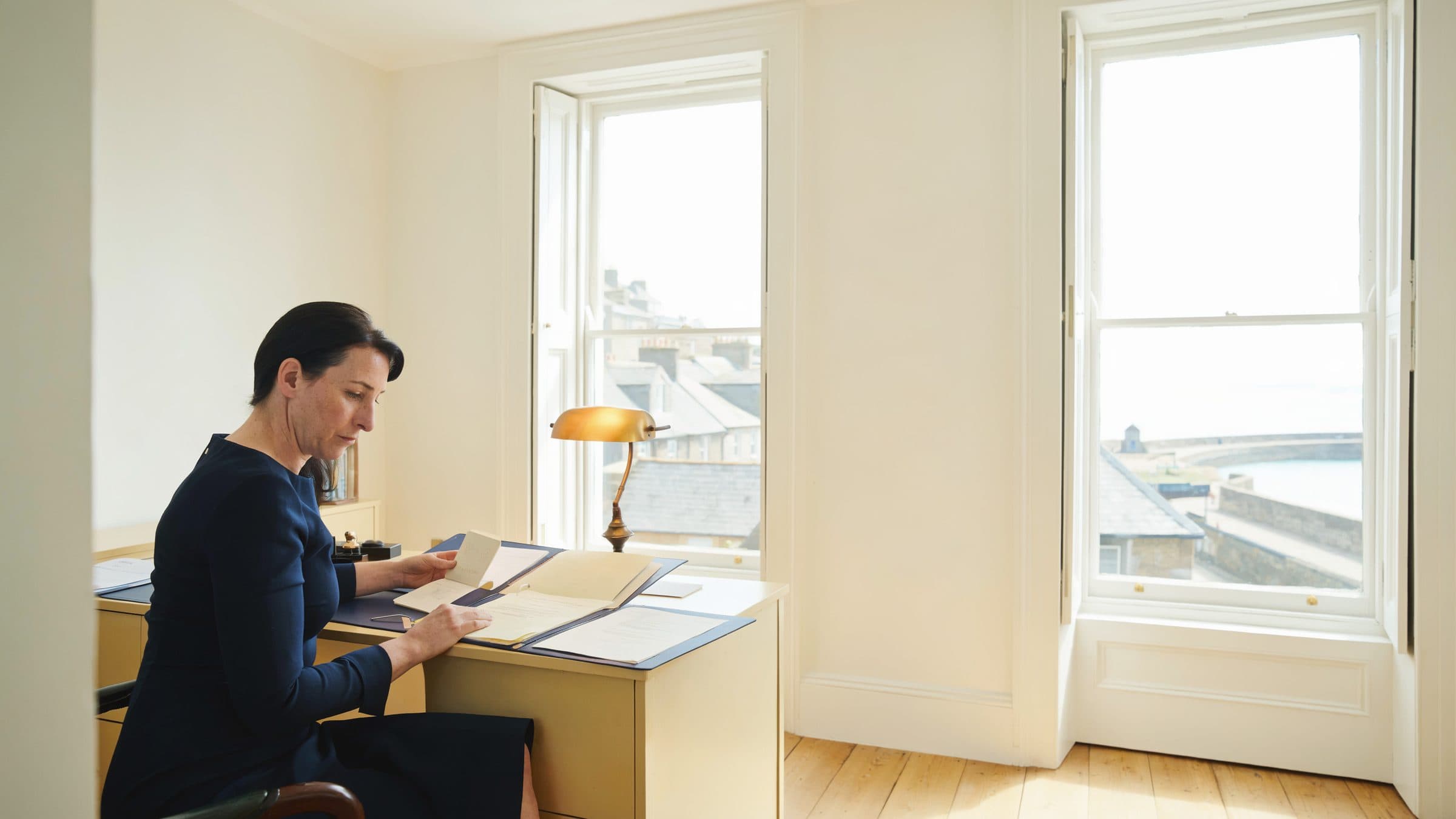 A compliance officer at a desk by a sash window, holding a passport beside an open client file, slate roofs and a harbour behind her.