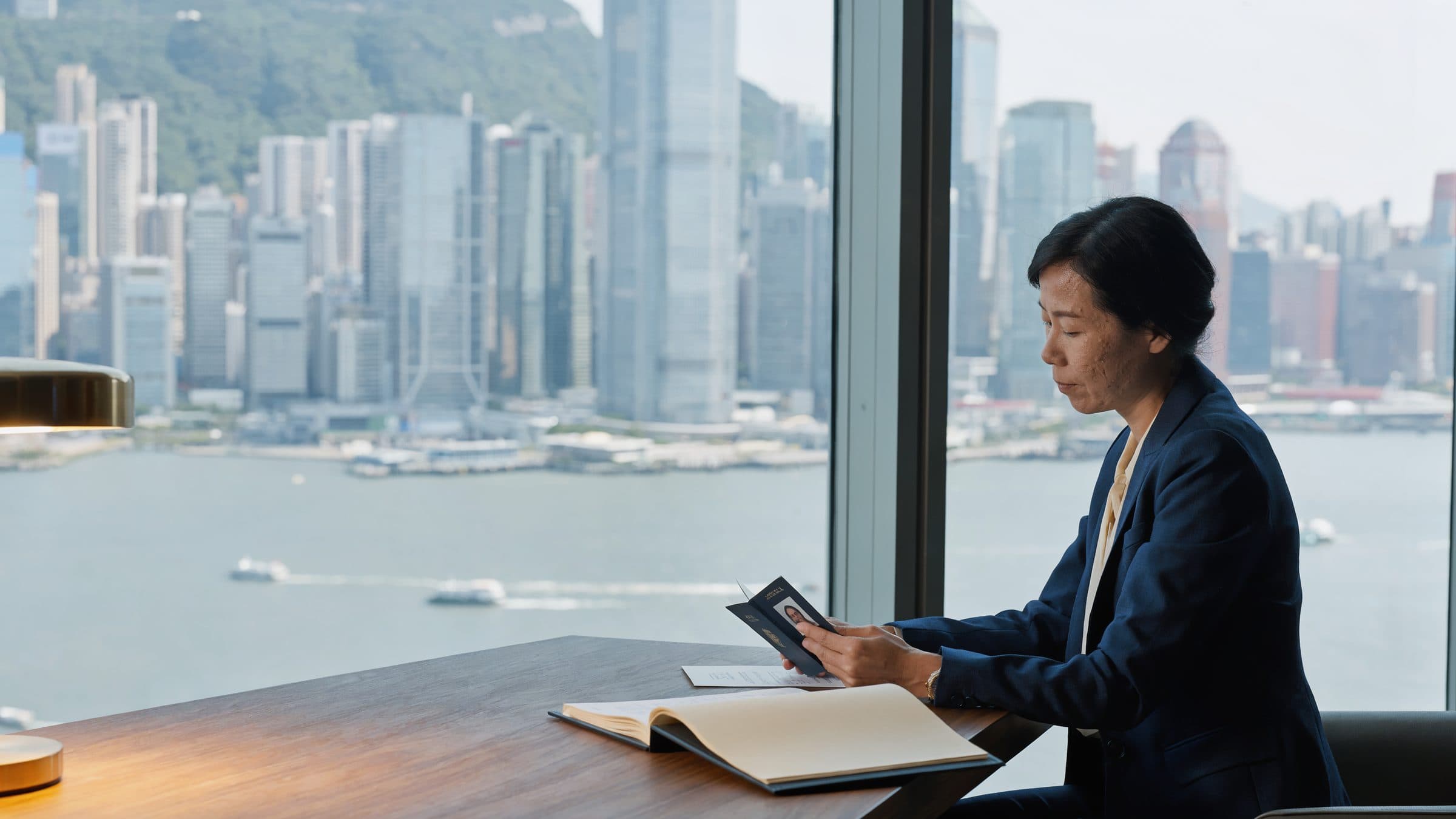 A compliance officer at a desk in a glass-walled office, a passport in her hand, a harbour and green hills behind her.