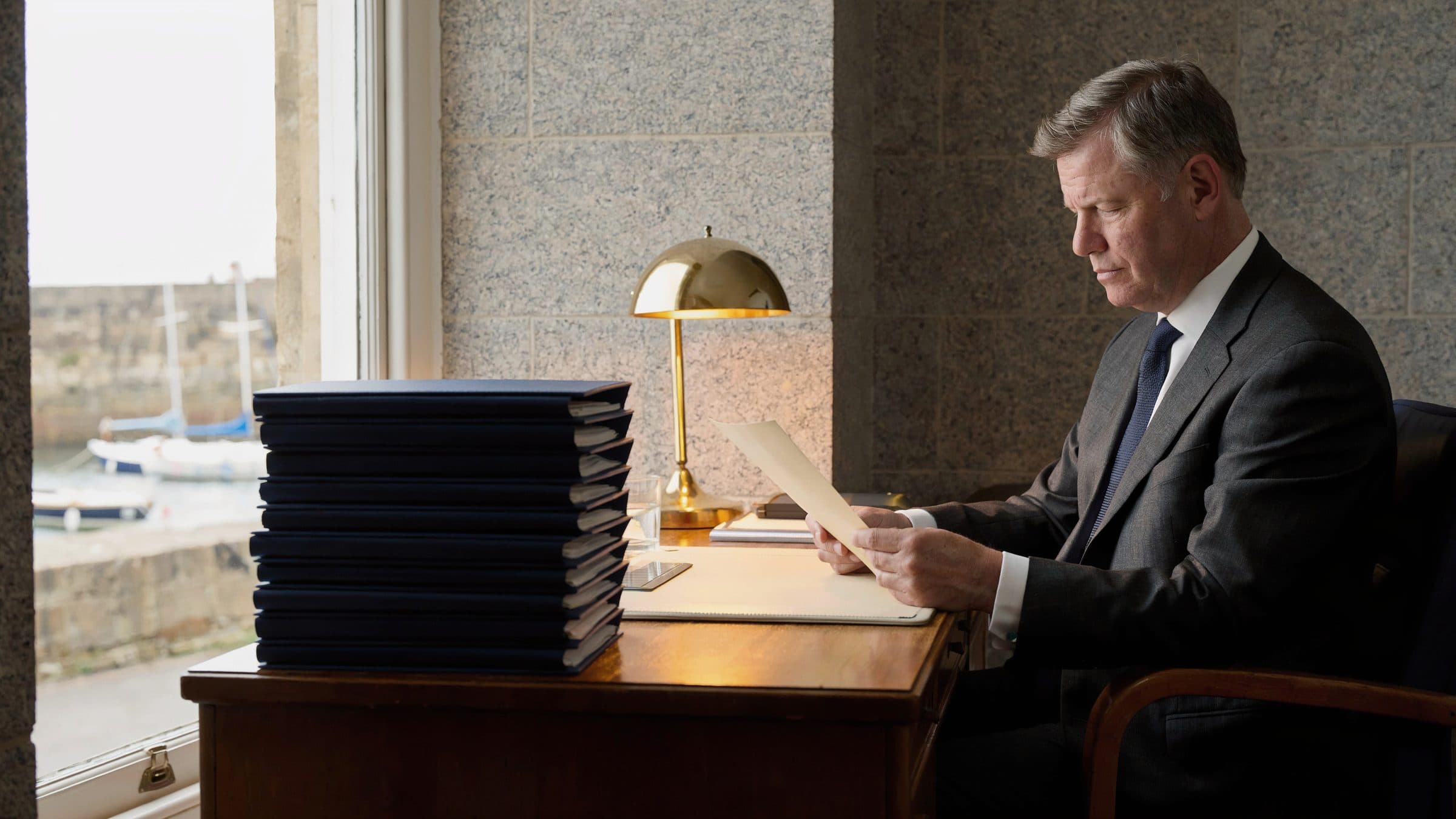 A trust officer reading a page at a desk with a stack of client files, a stone harbour of small boats through the window.