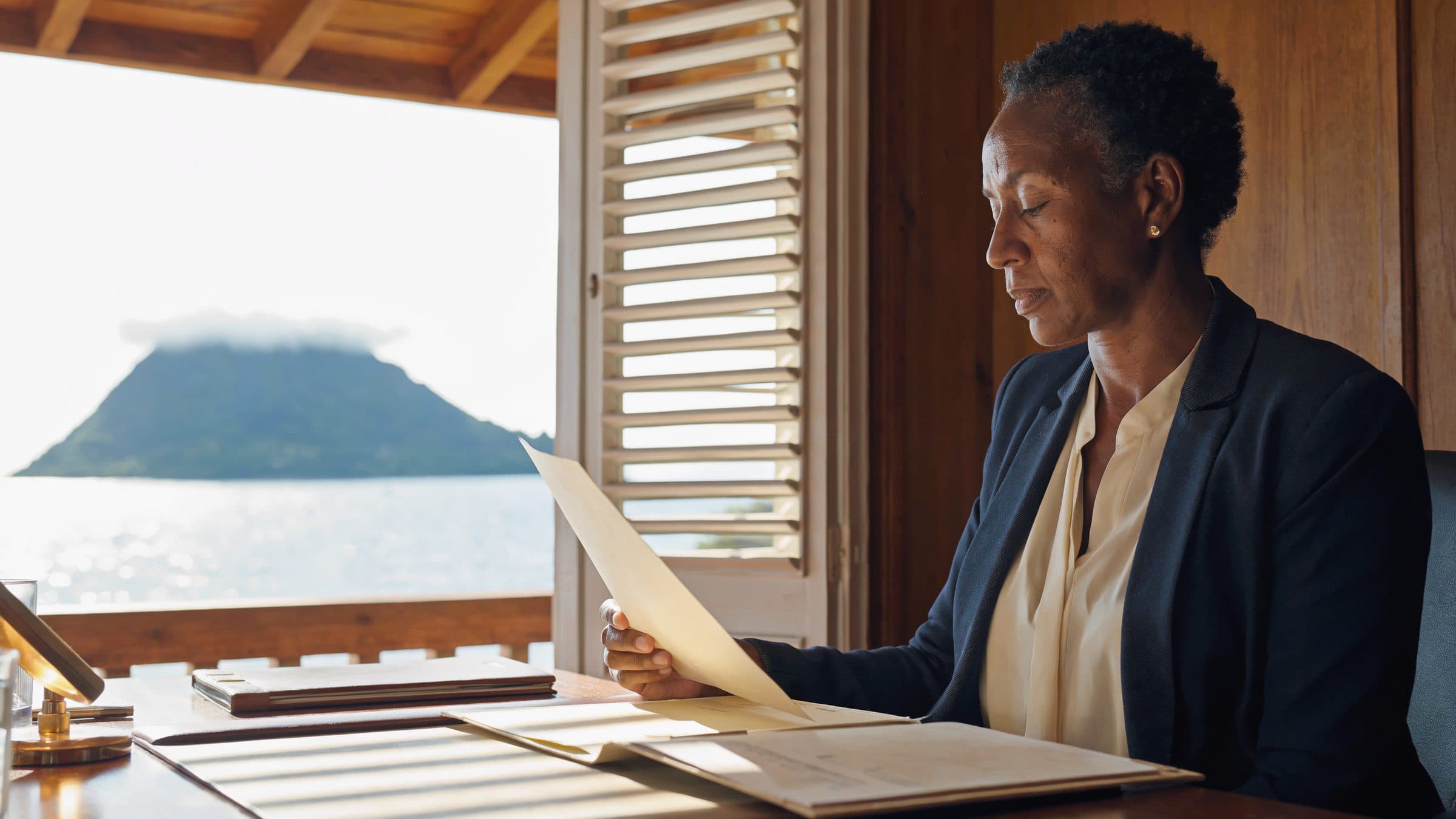 A trust officer reading a client file at a desk beside a louvred window, with a green peak across the water behind her.