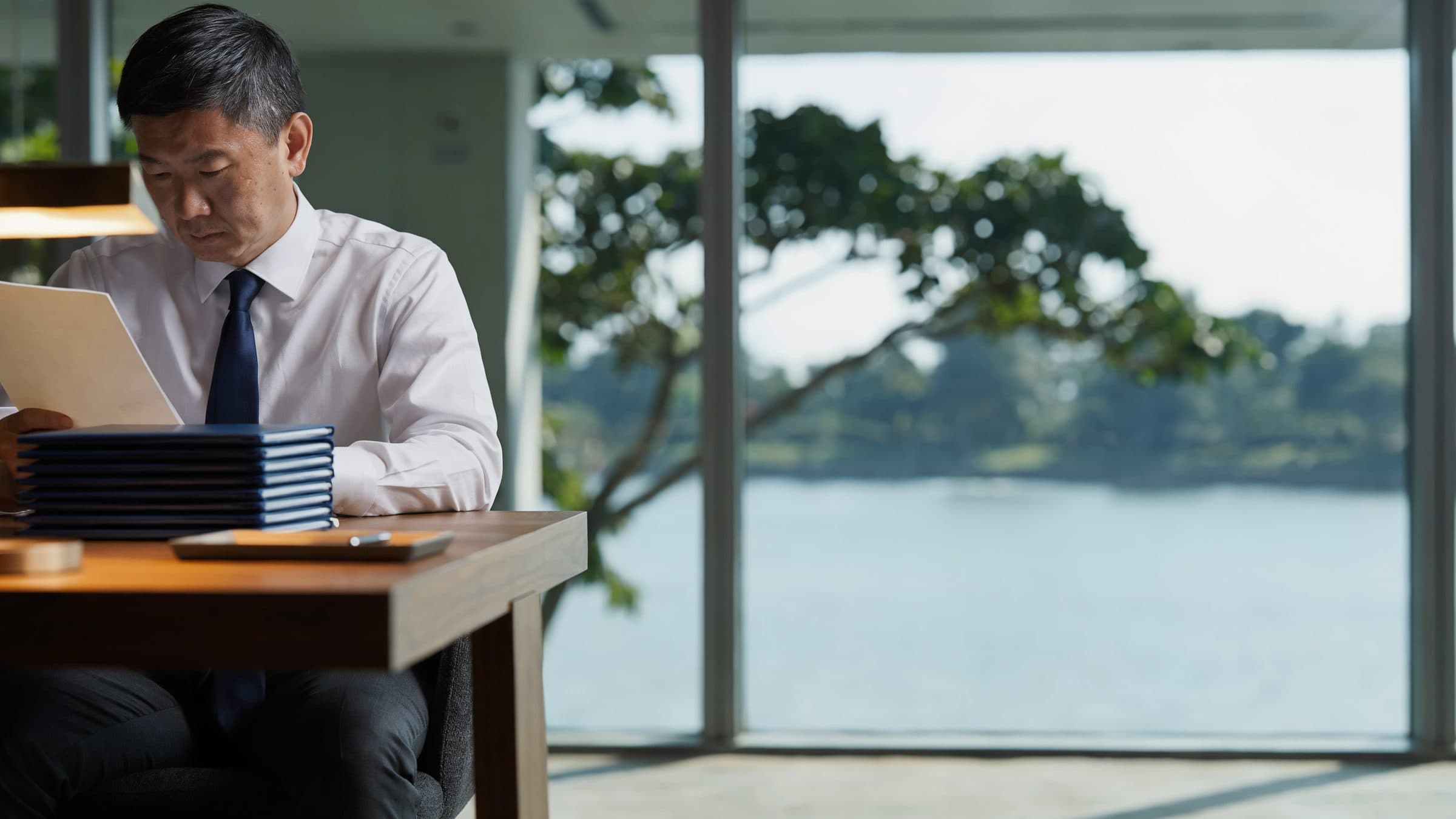 A corporate services officer reading a page at a desk with a stack of client files, a waterfront and garden trees through the window.