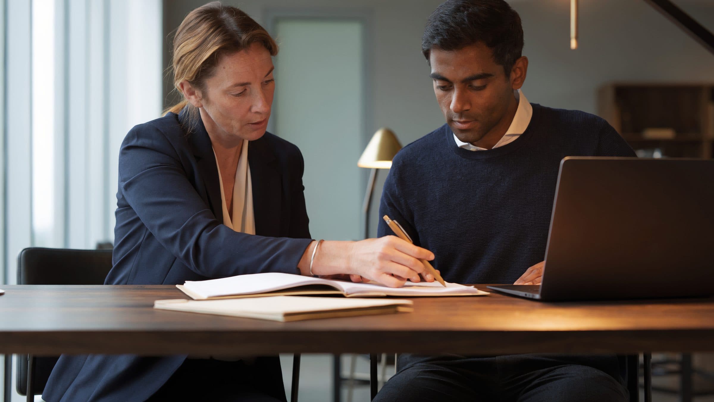 A compliance officer writing in an open file beside a developer with a laptop, at one desk.